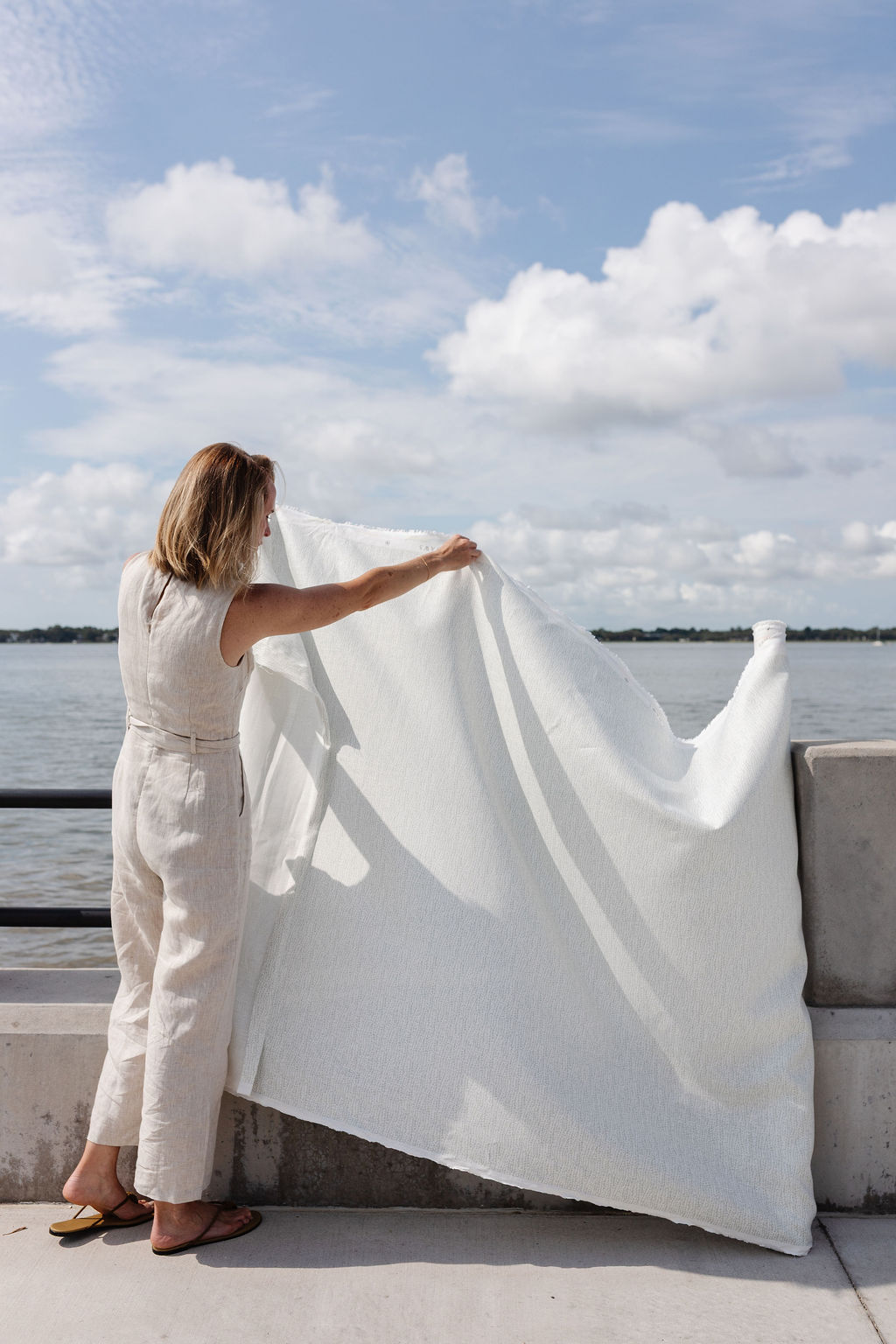 A woman stands by the water holding out a length of fabric in the breeze on a sunny day.