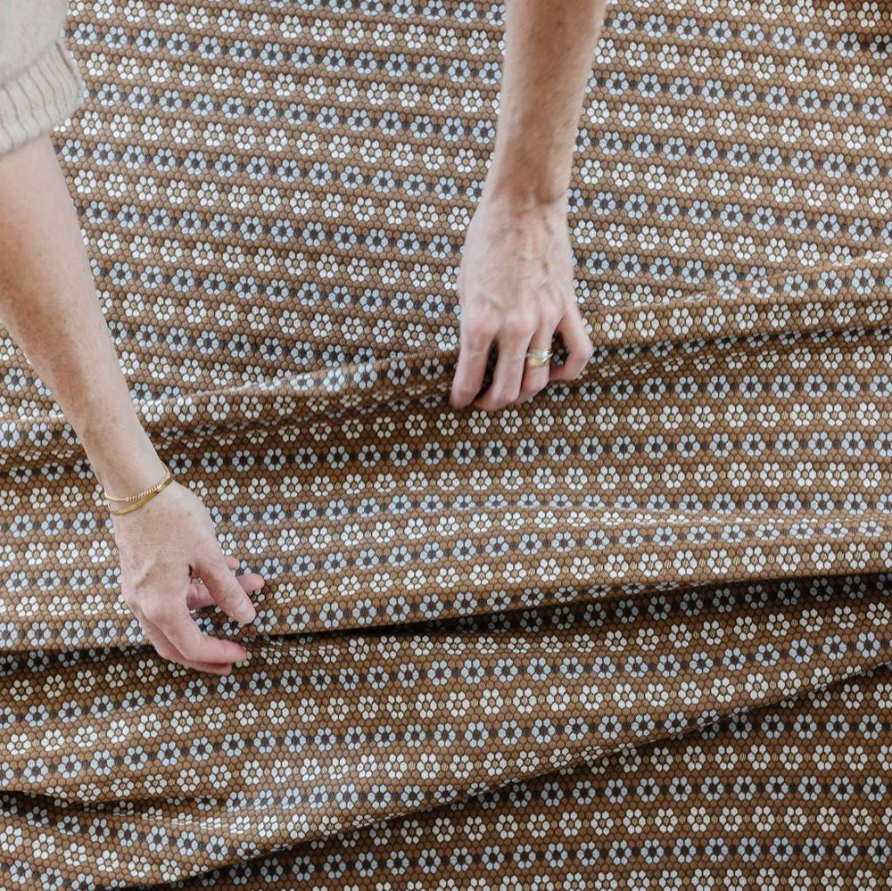 Hands smoothing a length of Emily Daws Nectar fabric in Walnut, a small floral woven pattern in brown with cream and soft blue accents.