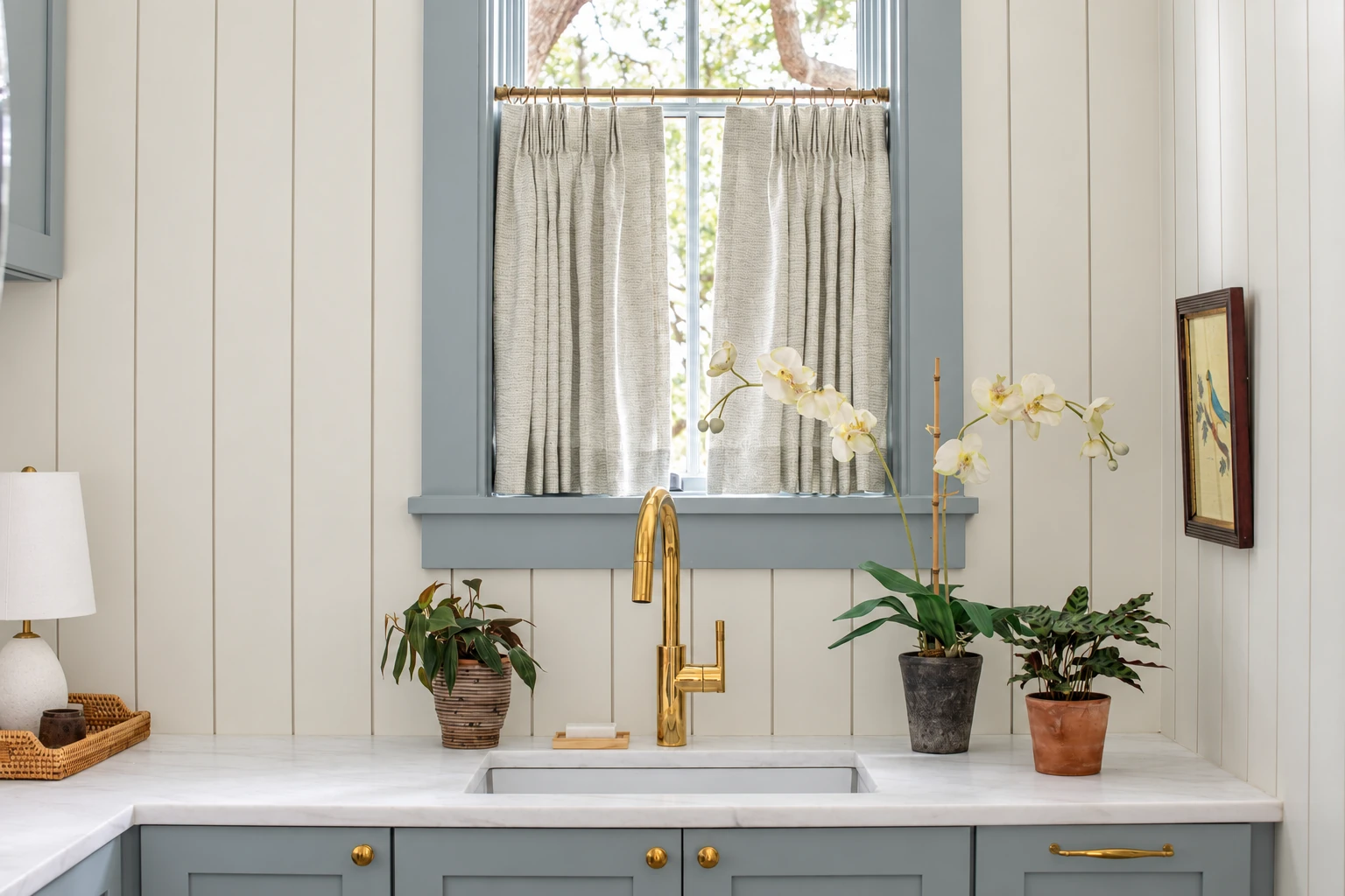 Emily Daws Harbor fabric in Celadon used as café curtains at a kitchen window with pale blue trim, white paneling and a brass faucet.