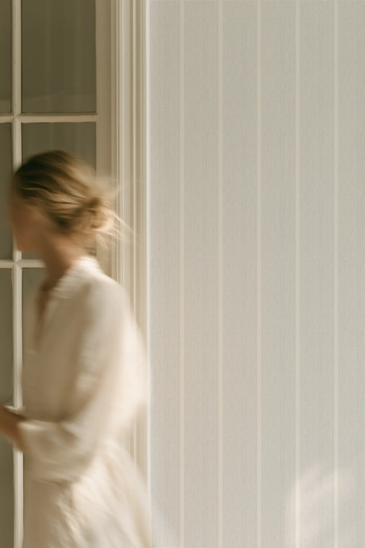 A blurred moving figure passes a glazed doorway beside the fine vertical stripes of Sandbar wallcovering in Sterling.