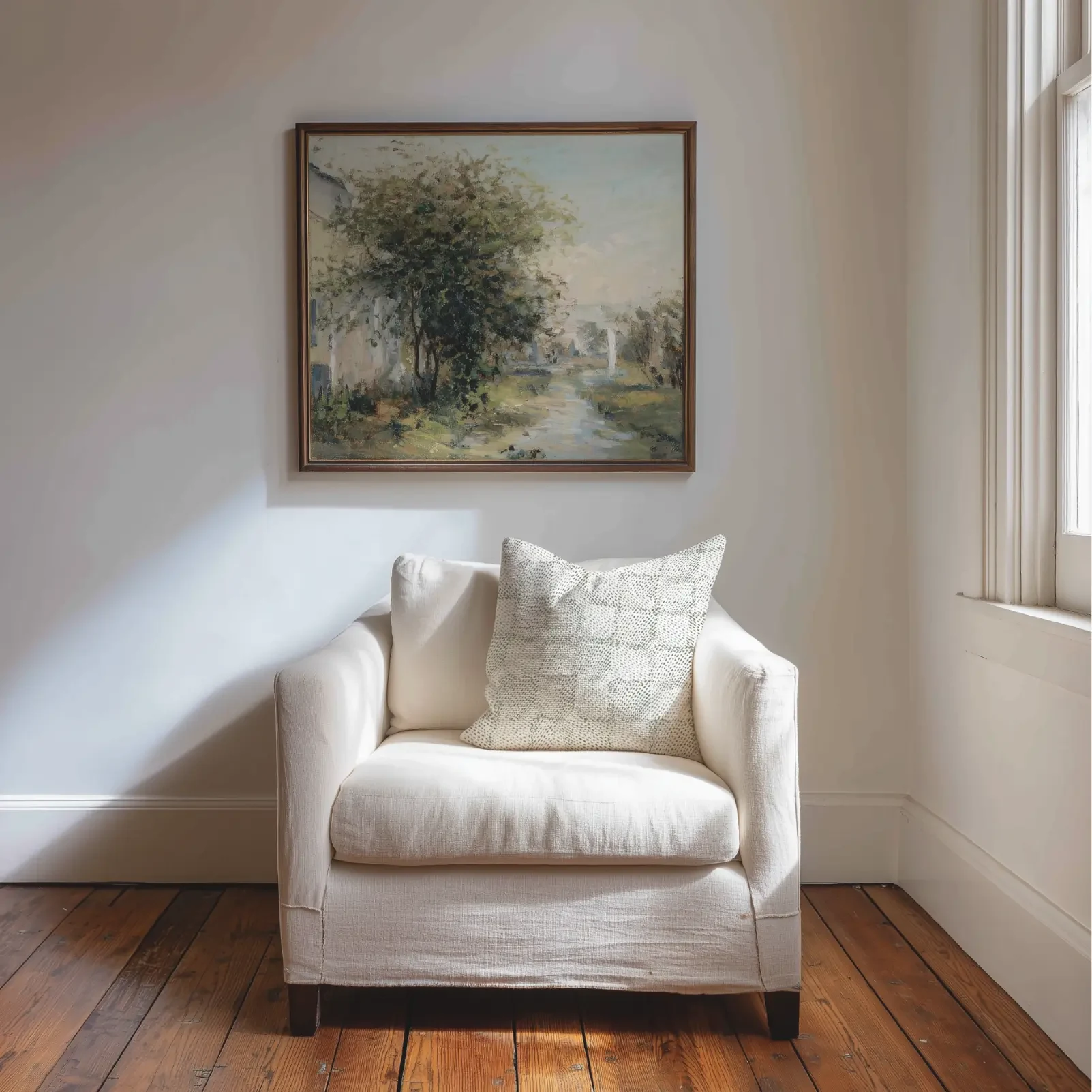 Pillow with a small dotted-check pattern resting on a white slipcovered armchair in a sunlit corner, with a small landscape painting on the wall above.
