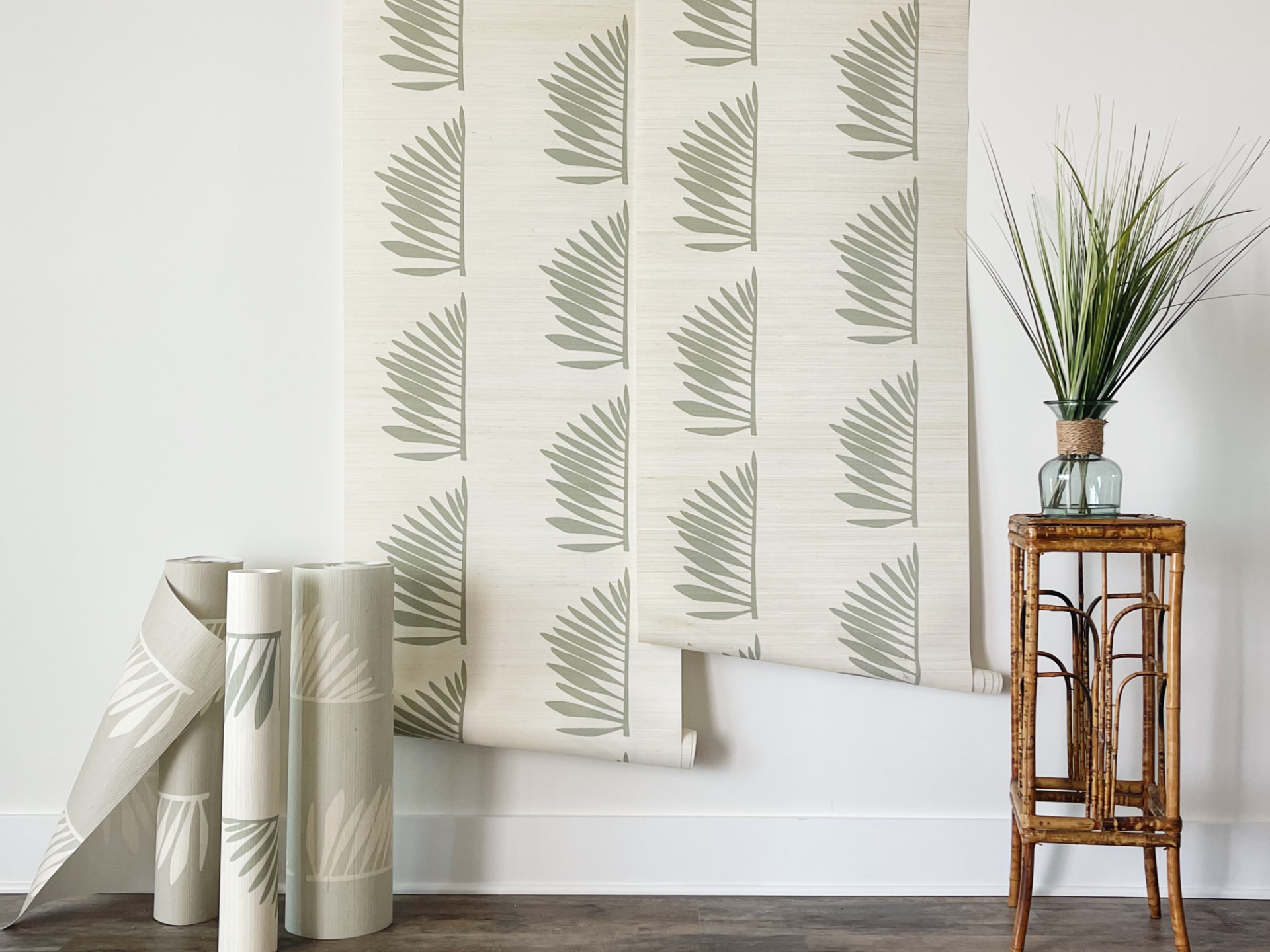 Palmetto Grasscloth in Sage shown as hanging wallpaper strips and rolls against a wall.