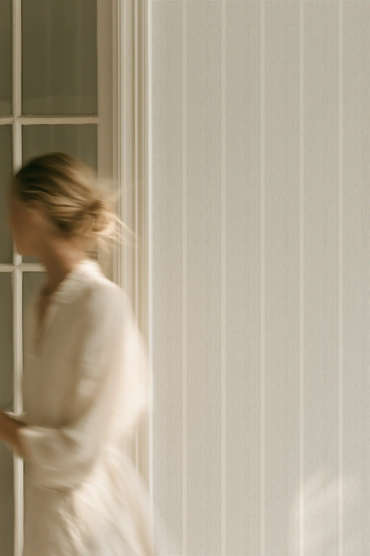A blurred moving figure beside a glazed doorway and the fine vertical stripes of Sandbar wallcovering in Driftwood.