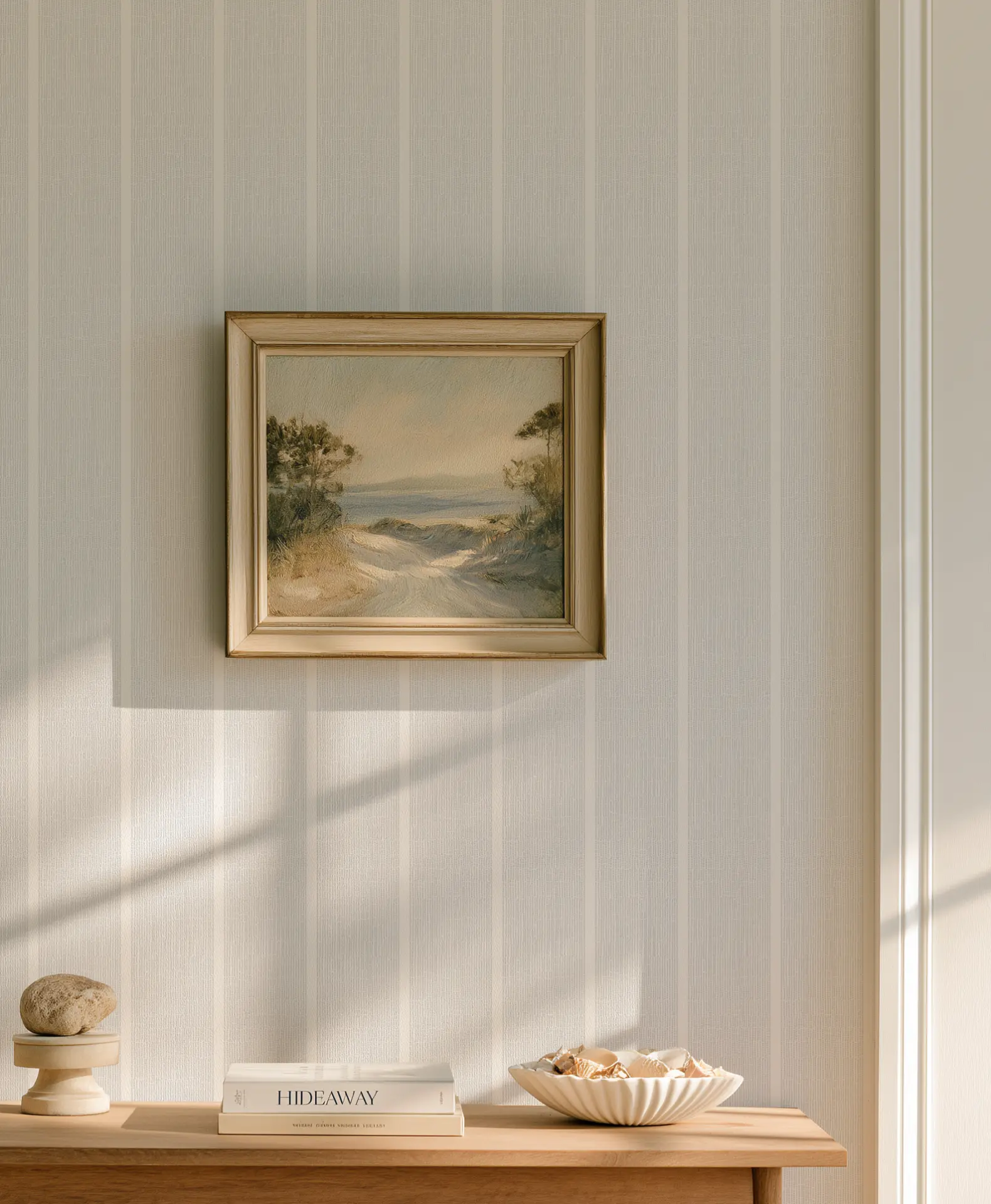 Sunlight falls across Sandbar wallcovering in Sterling above a wooden console with books, a bowl of shells, and framed coastal artwork.