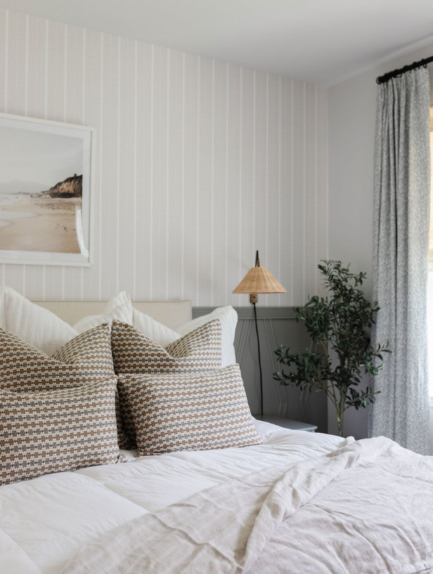 Emily Daws Nectar fabric in Raisin on layered bed pillows in a softly lit bedroom with striped walls and a rattan sconce.